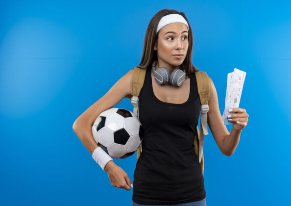 印象令人印象深刻的年轻漂亮的运动女孩戴着头带和腕带和背袋 脖子上戴着耳机 拿着机票 蓝色墙上有足球 还有复制空间球机票脖子