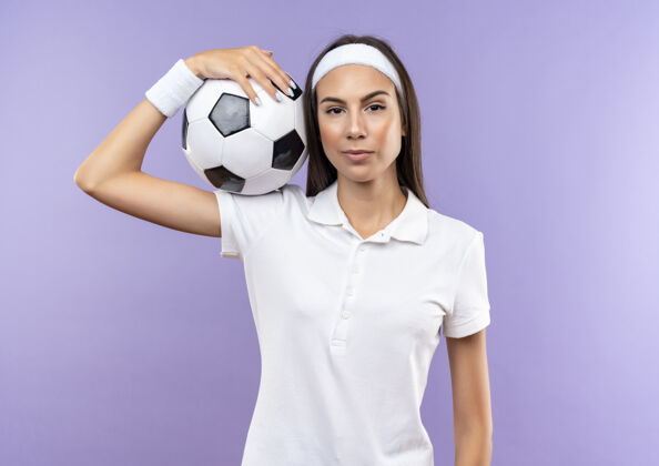 肩膀自信漂亮的运动女孩戴着头带和腕带肩上拿着足球隔离在紫色的墙上运动头带足球