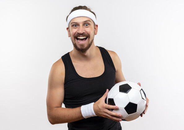 腕带快乐的年轻帅气的运动型男人戴着头带和手镯拿着足球看起来孤立在白墙上欢乐球头带
