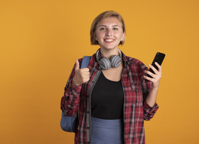 空间微笑着的年轻斯拉夫女学生戴着耳机 背着背包 拿着电话 竖起大拇指拇指耳机背包