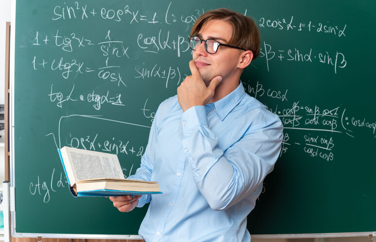边年轻的男老师戴着眼镜 拿着书 手放在下巴上 边看边思考 站在教室里的数学公式黑板旁男下巴近