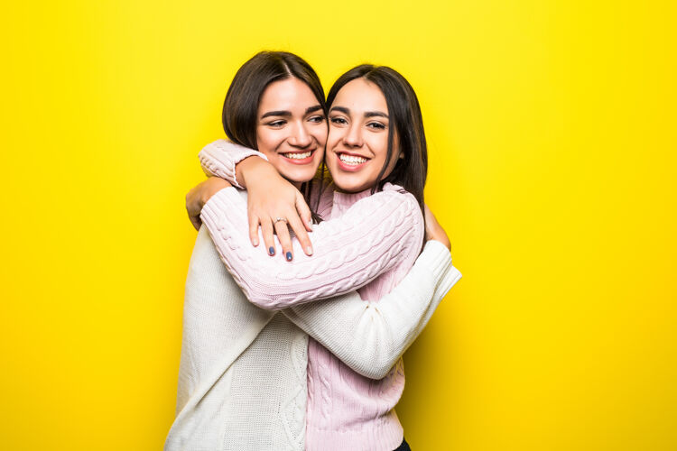 庆祝两个穿着毛衣的快乐女孩在黄色墙壁上拥抱的肖像人友谊人