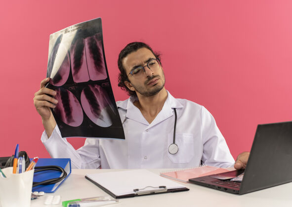X光年轻的男医生戴着医用眼镜 穿着医用长袍 听诊器坐在办公桌旁长袍工具空间