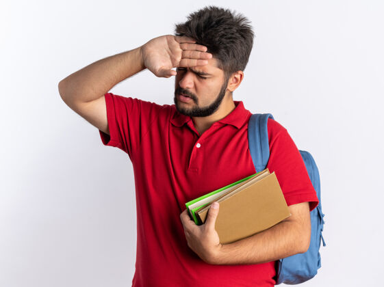 额头一个留着胡子的年轻学生 穿着红色马球衫 背着背包 拿着笔记本 看起来很不舒服 手放在额头上 站着发烧胡子小伙子红色