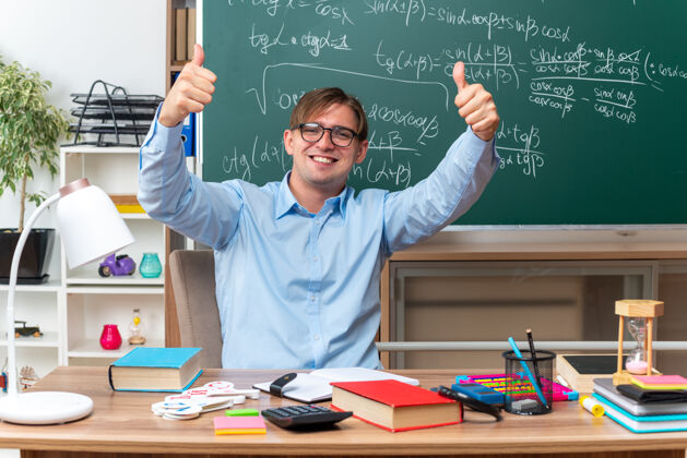 教室年轻的男老师戴着眼镜 面带微笑 竖起大拇指坐在课桌旁 教室的黑板前放着书和笔记穿前面男