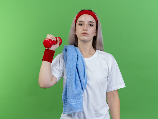 复制自信的年轻白种人运动女孩戴着背带戴着头带和腕带肩上带着毛巾拿着哑铃举行穿哑铃