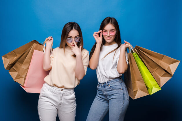 朋友快乐的两个女人 蓝色的墙上挂着五颜六色的购物袋兴奋微笑漂亮