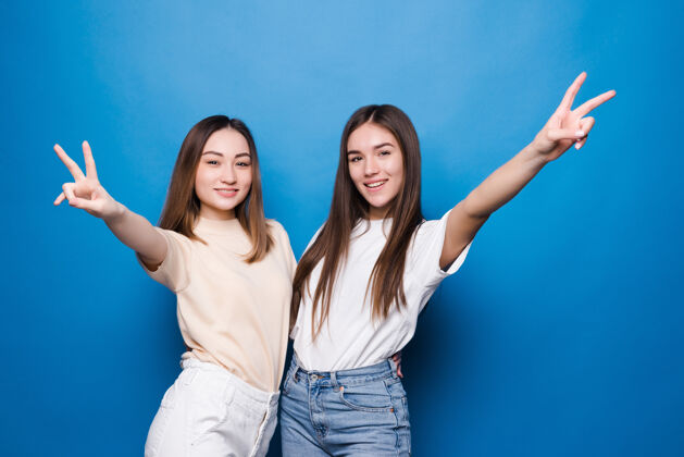 朋友年轻的两个女人伸出手指在蓝色的墙上做胜利的手势第二信心青少年正面