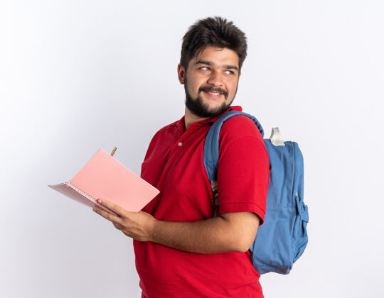 红色年轻的留着胡子的学生 穿着红色马球衫 背着书包 站在笔记本上写着自信的微笑胡须学生写作