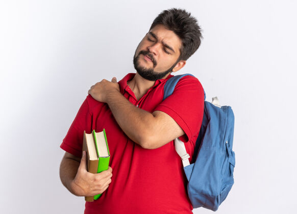 抱着年轻的留着胡子的学生 穿着红色马球衫 背着背包 拿着笔记本 看起来不舒服 摸着肩膀 站着感觉很痛触摸肩膀背包