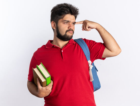 指着一个留着胡子的年轻学生 穿着红色马球衫 背着背包 拿着笔记本 用食指指着太阳穴 站在那里自信地思考着红色思考拿着