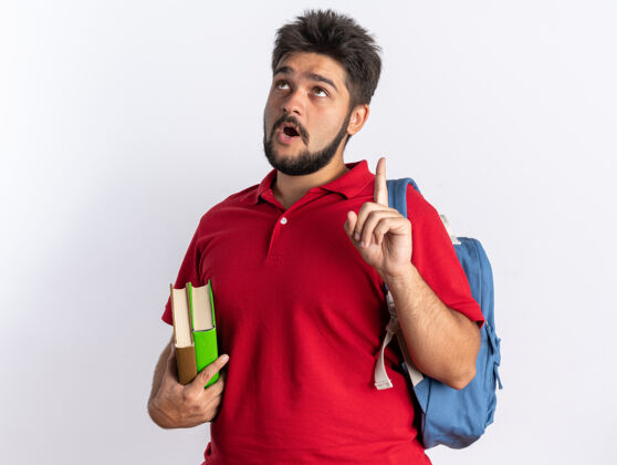 目录年轻的留着胡子的学生 穿着红色马球衫 背着书包 拿着笔记本 惊讶地抬起头 伸出食指 站着 有新的想法新年轻笔记本
