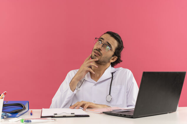 空间仰望着坐在办公桌旁的年轻男医生 戴着医用眼镜 穿着医用长袍 带着听诊器眼镜桌子穿