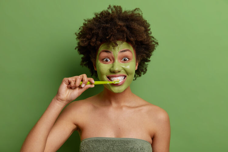 漂亮卷发的年轻女孩用非洲毛发刷牙进行日常卫生习惯 将面膜用在健康皮肤上 用毛巾包裹在鲜艳的绿色墙壁上头部口腔护理
