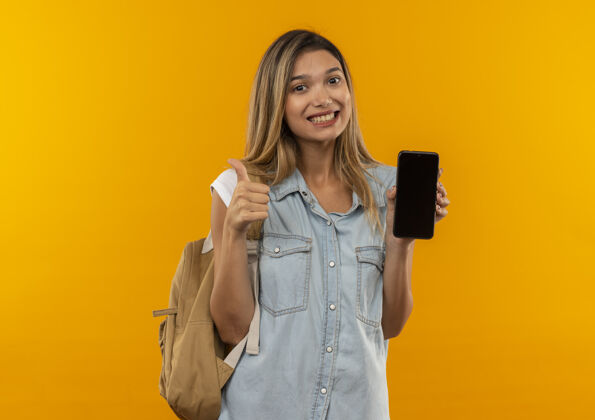 漂亮微笑着的年轻漂亮的女学生 背着书包 拿着手机 竖起大拇指 孤零零地站在橙色的墙上微笑衣服橙色
