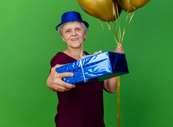 氦微笑的老妇人戴着聚会帽手持氦气球和礼品盒在绿色女人气球老人