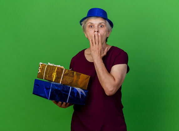 老人震惊的老妇人戴着聚会帽 手放在嘴上 手里拿着绿色的礼盒女人盒子帽子