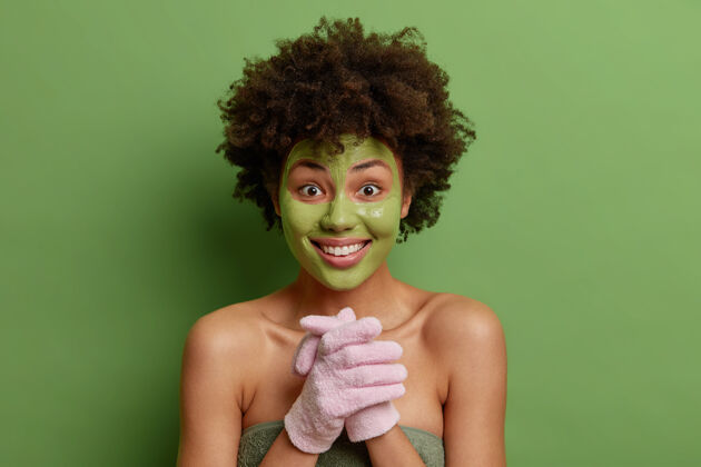 牙齿快乐卷曲的美国黑人女模特双手合十戴浴手套脸上敷绿色面膜焕发肌肤室内毛巾包姿势健康面膜滋养