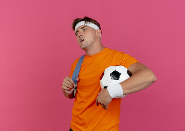 直体贴的年轻帅气的运动型男人戴着头带和腕带 背着一个包 手里拿着足球 背着一个包的带子 在粉色的墙上显得笔直孤立粉色腕带思想