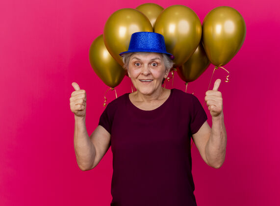 聚会戴着派对帽的快乐的老妇人站在氦气球前 竖起大拇指 两只手放在粉红色的气球上气球快乐氦