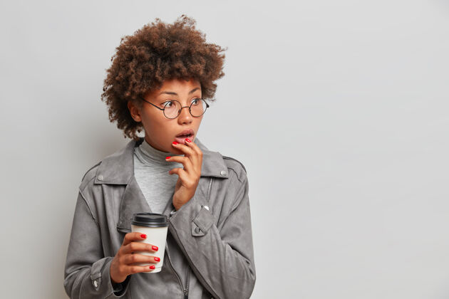 情绪惊讶的非裔美国妇女的肖像凝视着震惊的饮料外卖咖啡戴着圆眼镜时尚的夹克隔着灰色的墙休闲困惑害怕