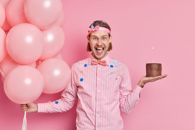 成人正面帅气的欧洲男人穿着衬衫 打着领结 手里拿着小巧克力蛋糕和一堆充气的气球 在粉红色的墙上享受生日聚会快乐乐观男孩