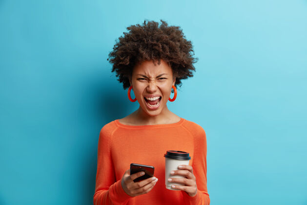 女性愤怒恼怒的女人用手机大声尖叫笑脸喝咖啡去穿橘色套头衫隔离在蓝色墙上做鬼脸后看到手机上有什么奇怪的东西不高兴肖像女人