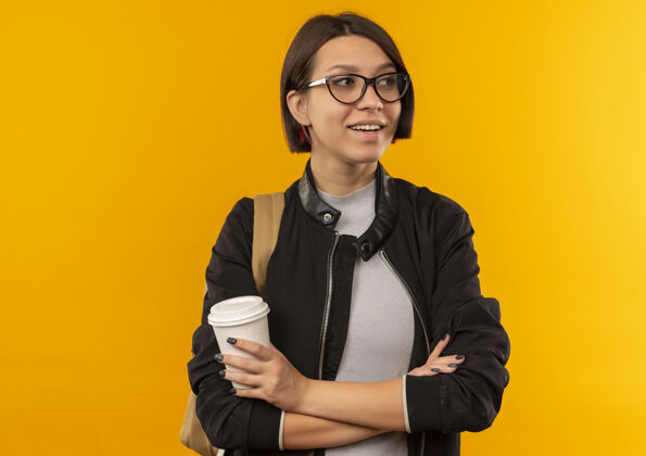 姿势面带微笑的年轻女学生戴着眼镜 背着书包 站着 抱着塑料咖啡杯 看着隔离在橙色墙上的一面脸抱着年轻