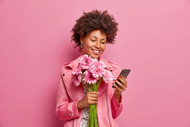 Swoosh高兴的非裔美国女士穿着时髦的衣服检查信息网上获得祝贺生日举行美丽的非洲菊花束有快乐的心情隔离在粉红色的墙上女性花店姿势