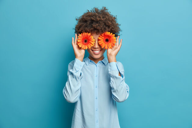人美丽开朗的女人 卷发遮住眼睛 拿着橙色非洲菊 穿着时髦的衬衫 隔着蓝色的墙积极的女花匠要为特别活动做装饰或花束花美丽快乐