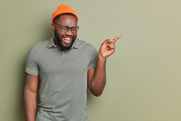 时尚快乐的美国黑人男子笑着开心地指着远处展示一些酷和有趣的东西戴着帽子和休闲t恤透明眼镜在演播室展示复制空间非洲年轻展示