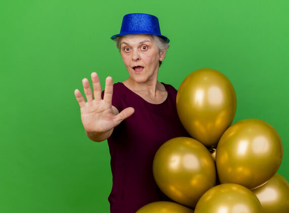 派对惊讶的老妇人戴着聚会帽站在那里 手里拿着氦气球 伸在绿色的草地上氦惊喜伸展