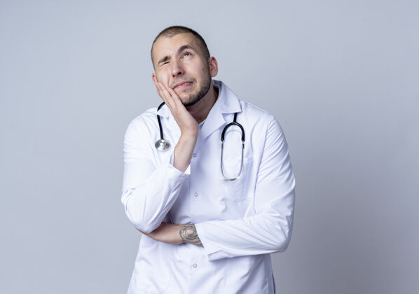 男性体贴的年轻男医生 穿着医用长袍 脖子上戴着听诊器 手放在脸颊上 一只眼睛闭着 仰望着白色的墙壁医学姿势听诊器