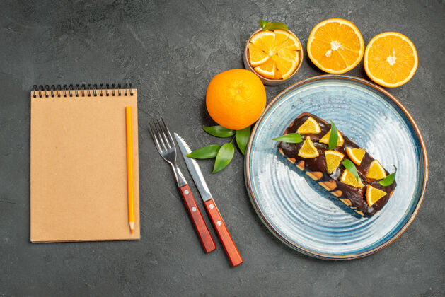 切割黄色的整个柠檬和切好吃的蛋糕用刀叉旁边的笔记本在黑暗的桌子上饮食深色画笔