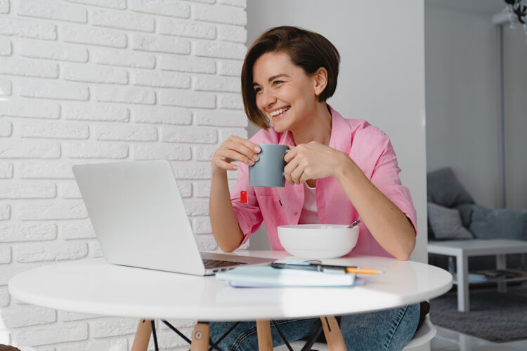 电脑穿着粉色衬衫的坦率微笑的女人在家里吃早餐 在桌旁用笔记本电脑上网 在家里吃麦片工作学习女性