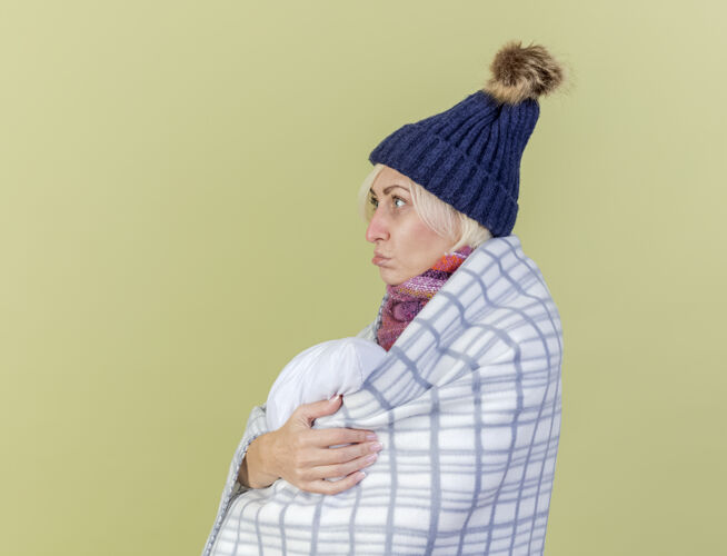冬天悲伤的年轻金发病女人戴着冬天的帽子和围巾包裹在格子布站在一边 拥抱着橄榄绿墙上的枕头隔离人站着脸