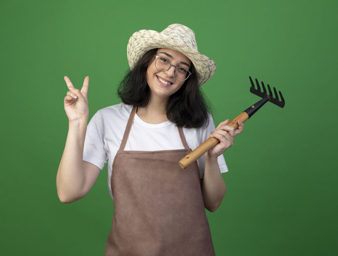 公民微笑着的年轻黑发女园丁戴着眼镜和制服戴着园艺帽拿着耙子和手势胜利的手势孤立在绿色的墙上制服眼镜拿着