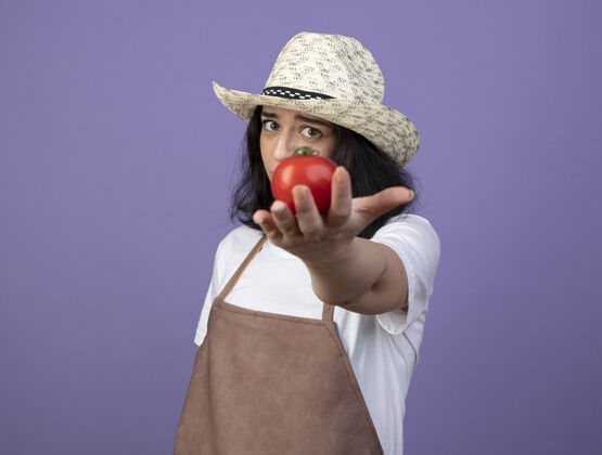 帽子失望的年轻黑发女园丁穿着制服 戴着园艺帽 在紫色的墙上伸出番茄花园西红柿人