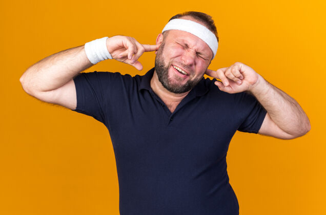 男人恼怒的成年斯拉夫运动男子戴着头带和腕带闭上耳朵手指孤立在橙色墙上的复制空间手指腕带斯拉夫人