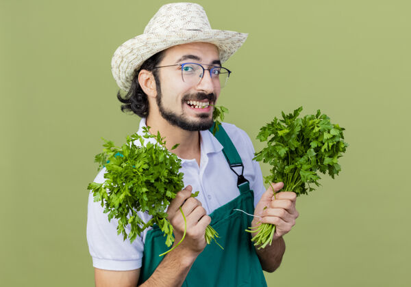 快乐年轻的留着胡须的园丁 穿着连体衣 戴着帽子 手里拿着新鲜的草药 站在浅绿色的墙上 面带微笑地看着前面胡须表情站着