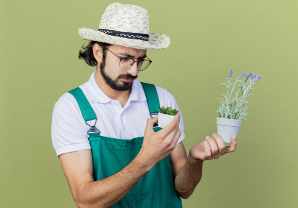 姿势年轻的留着胡子的园丁 穿着连体衣 戴着帽子 手里拿着盆栽植物 站在浅绿色的墙上 带着悲伤的表情看着它们帽子人连身衣