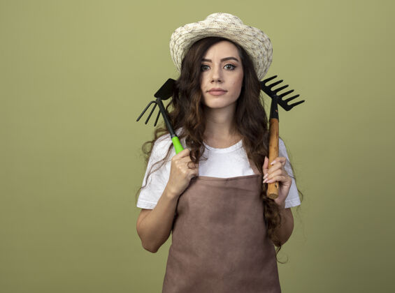 表情自信的年轻女园丁穿着制服 戴着园艺帽 把耙子和锄头耙子放在隔离在橄榄绿墙上的肩膀上年轻锄头脸