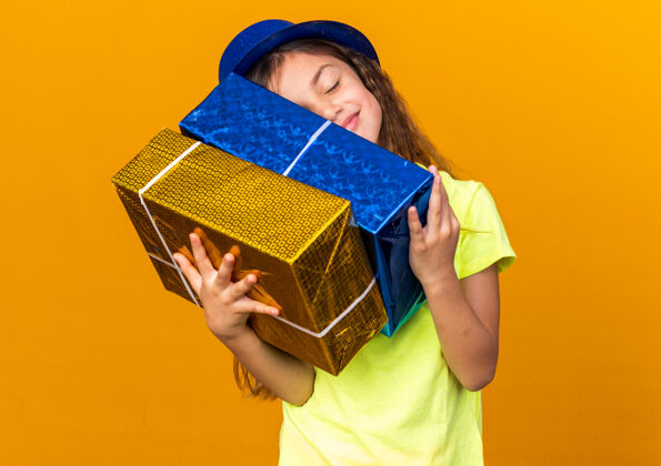 头可爱的白人小女孩戴着蓝色派对帽 把头戴在礼品盒上 隔离在橙色的墙上 留着复印空间孤立的抱着帽子