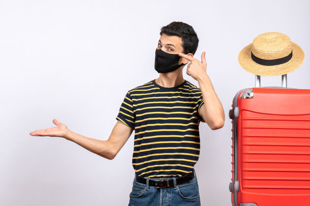 黑色正面图：戴着黑色面具的年轻游客站在红色手提箱旁 展示着一些东西男人红色显示