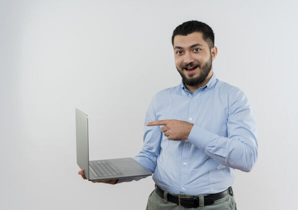 目录身穿蓝色衬衫 留着胡须的年轻人拿着笔记本电脑 用食指指着笔记本电脑 站在白色的墙上 开心而积极地微笑着微笑人表情