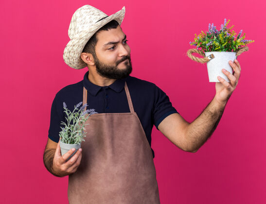 年轻困惑的年轻白人男园丁戴着园艺帽拿着花盆看着粉色墙壁上的复制空间困惑男性帽子