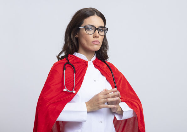 公民身着医生制服 戴着红色斗篷 戴着眼镜的听诊器 自信的女超人在白色的墙壁上双手合十表情人斗篷