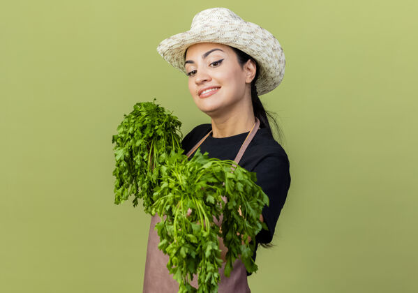 新鲜年轻漂亮的女园丁 围着围裙 戴着帽子 手里拿着新鲜的药草 站在浅绿的墙上 面带微笑地看着前面花园美丽表情
