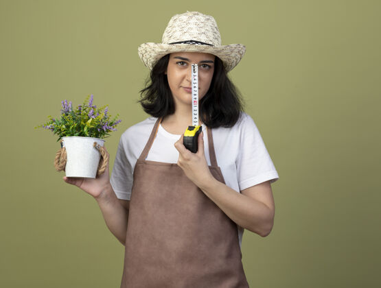 胶带自信的年轻黑发女园丁穿着制服 戴着园艺帽 把花盆和卷尺隔离在橄榄绿的墙上测量表情持有