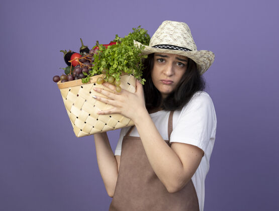 帽子失望的年轻黑发女园丁穿着制服 戴着园艺帽 把菜篮子孤立地放在紫色的墙上制服穿着篮子